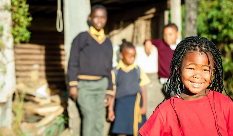 A pretty african girl with braided hair and a bright red shirt smiling confidently with her siblings in the backround watching over her. Photo: Nolte Lourens - stock.adobe.com